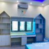 Hình ảnh thực tế chiếc bàn đôi dành cho bé trai và bé gái với màu gỗ vàng nâu cánh trắng, bên còn lại màu xanh, thiết kế ôm cửa sổ, đỉnh của giá sách hình mái nhà rất đẹp mắt